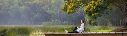 Panorama of asian woman sitting on the edge of dock with peaceful natural park during summer with yellow flower blossom for serene and relaxation outdoor recreation