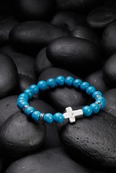 Detailed shot of blue bracelet made of turquoise beads with veins and decorated with silver charms and ivory stone cross. The stylish bracelet is located on black pebble stones. 