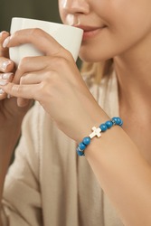 Cropped shot of a lady with turquoise beaded bracelet on her wrist. The bracelet is decorated with ivory stone cross and silver charms. The girl in beige blouse is holding white cup in her hands.     