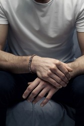  Cropped shot of a man with beaded bracelet made of smoky gray stone and decorated with blue cat eye bead. Man in white t-shirt and dark jeans is sitting with crossed hands.                           