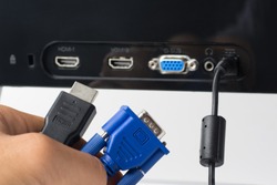 Man`s hand holds HDMI and VGA cables against a monitor with ports. Choise between modern HDMI and old VGA connection