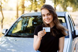 Attractive young woman wearing black T-shirt standing in front of modern car bragging about receiving driving license showing licence to the camera and feeling happy