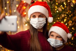 Mother and son make selfie near the fir-tree, both of them in christmas hats and medical masks. Winter holidays celebration during quarantine.