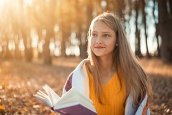 Beautiful girl reading a book and dreaming of something positive. Smiling blond girl likes to read books outdoors in autumn park