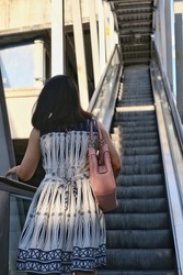 The back view of an Asian woman in white and blue dress is going up an escalator to a sky train station in the morning.  