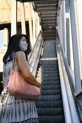 The back view of an Asian woman with a white face mask, wearing a white and blue dress is going up an escalator to a sky train station in the morning.  