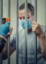An infrared thermometer is pointed to a prisoner's forehead. A middle-aged man in mask stands behind bars of the cell