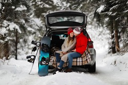 A young couple of snowboarders man and woman are sitting in the trunk of his car in an hugs. A walk of a young couple in the winter pine forest. Winter active leisure.