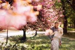 Beautiful, cheerful and lively newlyweds, groom and bride are hugging near the blooming pink cherry blossom. Wedding portrait of a close-up of a smiling bearded groom and a cute bride