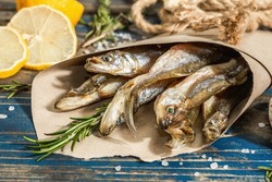 Sun-dried smelt in a paper package. Salted fish with marine decor. Lemon, fresh rosemary, sea rope. Blue nautical wooden background, close up