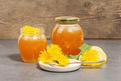 Dandelion jam or honey in the glass jar with fresh blooming dandelion flowers. Seasonal product, trendy hard light, dark shadow. Stone concrete background, close up