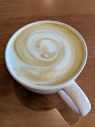 White cup of Cappuccino with swirl in foam on saucer on table.
