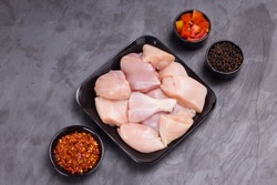 Raw chicken tender curry cut without skin arranged on black plate and garnished with tomato slices, chilli flakes and pepper on stone textured or graphite colour background