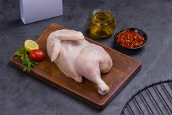 Raw half chicken with skin arranged on brown wooden board and garnished with parsely,small tomato,chilli flakes ,oil and lemon slices with delivery box on stone textured background,isolated