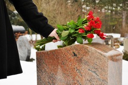 Woman in a snowy graveyard in winter puts a bouquet of roses on a tombstone of grave