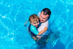 Father playing with child in a swimming pool. Dad embracing and protecting  his son in a blue water. Toddler affraid of water.