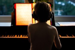 Boy in headphones sitting at the piano in the evening, view from the back. Child practicing play pianoat home, music education concept.