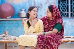 Cheerful rural Indian mother and young child daughter laughing having a good time together, sitting on traditional bed, Cute adorable girl kid in braided hair and mum in red sari, happy parent bonding