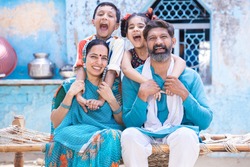 Cheerful rural indian family  sitting on traditional bed at village home, excited little kids having fun with parents.  daughter and son hug father and mother. happy life, playful children. 