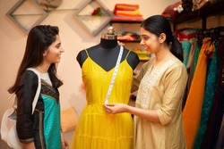 Young happy indian woman,tailor seamstress,smiling fashion designer showing customer or client dress measurements with tape,measuring on mannequin,standing in garment workshop,atelier or studio.
