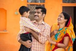 Young happy indian parents holding cute little child having fun and laughing while standing outdoor at village house. Mustache man wearing kurta and woman wear sari. rural india concept.