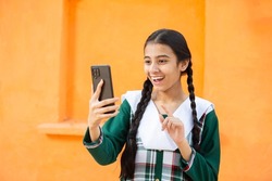 Young Happy Excited Rural Indian girl do video call on Smart phone, Cheerful braided female using digital android mobile phone or cellphone device against orange background, skill india.