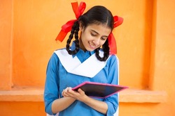 Young beautiful happy indian school girl write on slate against orange background, Smiling braided hair female teenager kid study with black board. education concept. rural india.