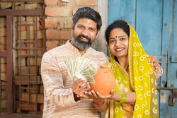 Portrait of happy rural couple holding indian rupee notes and clay money box or gullak. traditional piggy bank, save money, Investment and banking