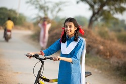 Portrait of happy young rural indian girl wearing blue school uniform standing with bicycle at village street.