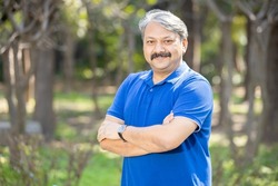 Happy senior indian asian man with arms crossed standing at park outdoor, Old mustache and grey haired male smiling wearing blue t-shirt. Copy space.