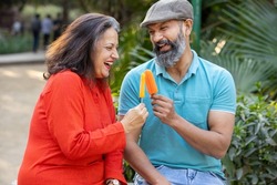 Happy indian senior couple eating ice lolly or ice cream in a park outdoor, old mature people enjoy retirement life. summer holidays.