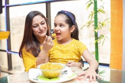 Young Indian mother feed her little daughter noodles breakfast at table, Little girl child having food with her mom spoon feeding . selective focus