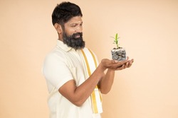 Happy South Indian man holding jar filled with money saving coins and plant growing isolated on beige background. Investment And Interest Concept