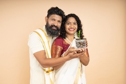 Happy South Indian couple holding jar filled with money saving coins and plant growing isolated on beige background. Investment And Interest Concept