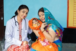 Portrait of Young indian pediatrician doctor sitting with Mother and her new born baby looking at camera, Rural healthcare camp concept.
