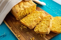 Cornbread is bread containing cornmeal, corn flour. Beautiful homemade yellow sliced cornbread loaf under cotton towel. Blue wood background. Above view.