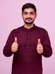 Young indian man wearing kurta standing over isolated background doing happy thumbs up gesture with hand. Approving expression looking at the camera showing success.
