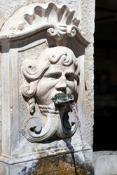 The antique old stone fountain with drinking water in the north of Italy, Lombardy, Brescia. Head of a man with long hair and a metal frog in his mouth a tap for water. Western Europe. Heritage.