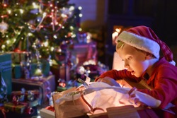 Lovely little boy with a santa claus hat opens a gift in front of the Christmas tree lit up, in the warm atmosphere of Christmas,  a wood stove in the background