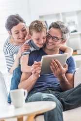 At home, Dad, mom and their young son having fun by gaming together on a tablet, they are sitting on a white couch in the living room and the boy looks at the screen over the shoulder of his father