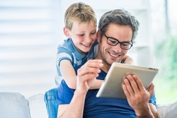 At home, a father and his young son having fun by gaming on a tablet, dad sits on a white couch and the boy looks at the screen over the shoulder of his father and giving him advices to win