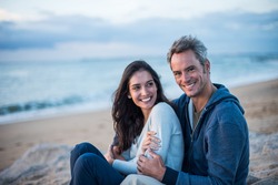Beautiful couple sitting on a rock at the beach watching the sunset. They wear casual clothes