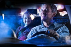 front view. A handsome man driving his car at night in the rain.  At the back his little daughter smiles to her dad in the mirror