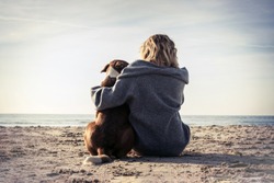 Young woman sitting and hugging dog on the beach. Friendship concept - woman and dog sitting together on a beach and enjoying sunrise. Back view colorized image