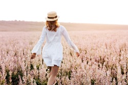 Back view of a pretty young woman in white dress holding straw hat running walking at the sage flower field. Beautiful girl enjoying a field of flowers, relaxing outdoors, having fun, harmony concept.