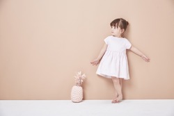 The little girl stood in front of the wall in a white dress, lively and lovely.