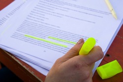 Female hand holding highlighter pen, highlighting / marking important texts in study sheets on table background.
