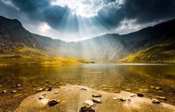 The sun breaks through the clouds over a mountain lake. Sun breaking through clouds over mountain lake landscape. Mountain lake landscape. Lake in mountains