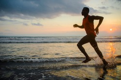silhouette of young athletic and fit african american sport man doing running workout on sunset at the beach training hard jogging barefoot in healthy lifestyle concept
