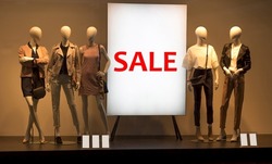 Five mannequins standing in store window display of women's casual clothing near the stand. Sale